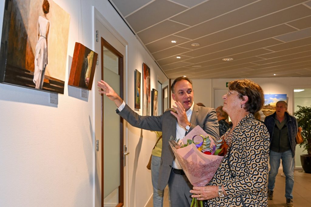 Een vrouw draagt een bos bloemen in haar handen, terwijl ze met wethouder Philippe van Ham praat over een schilderij.