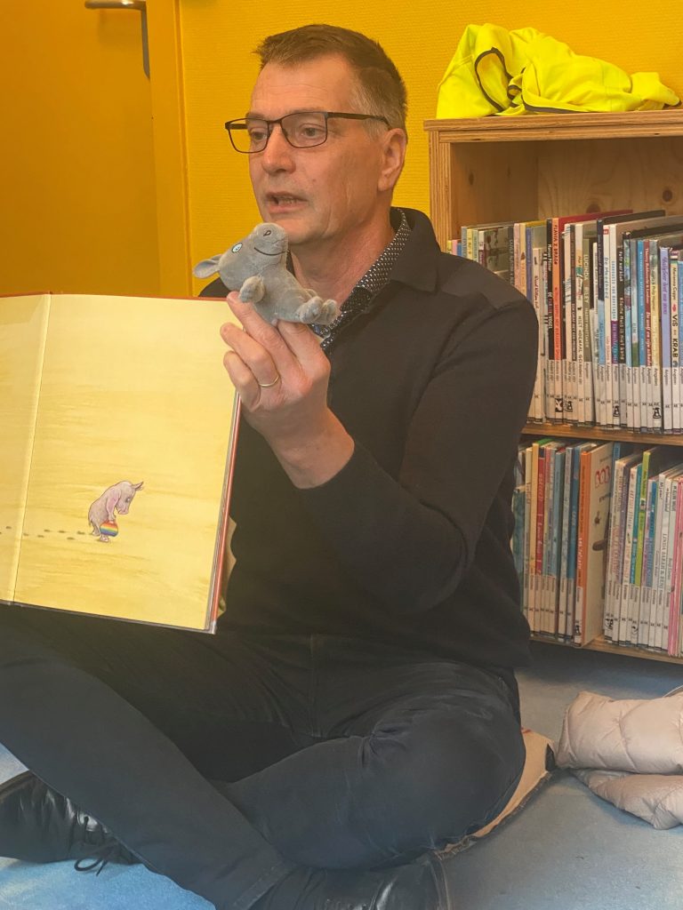 wethouder Win Bijman zit op de grond in een bibliotheek en laat een pagina van het prentenboek 'Rinus' zien.