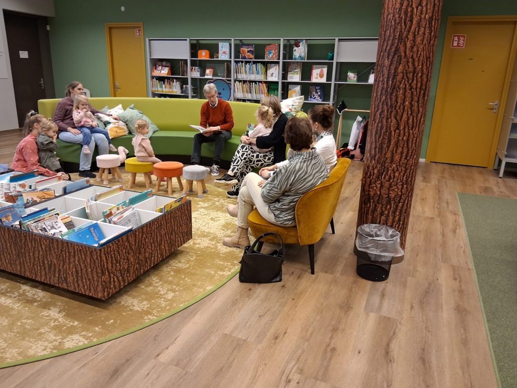 Wethouder Sjon Wagenaar zit in een bibliotheek en heeft het prentenboek 'Rinus' op schoot. Kinderen zitten bij ouderen op schoot en luisteren naar het verhaal.