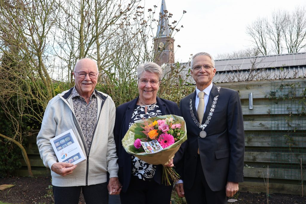 Meneer en mevrouw Dudink-Tames staan met burgemeester Pieter Dijkman in de tuin. Op de achtergrond zie je een schutting, bomen en een kerk.