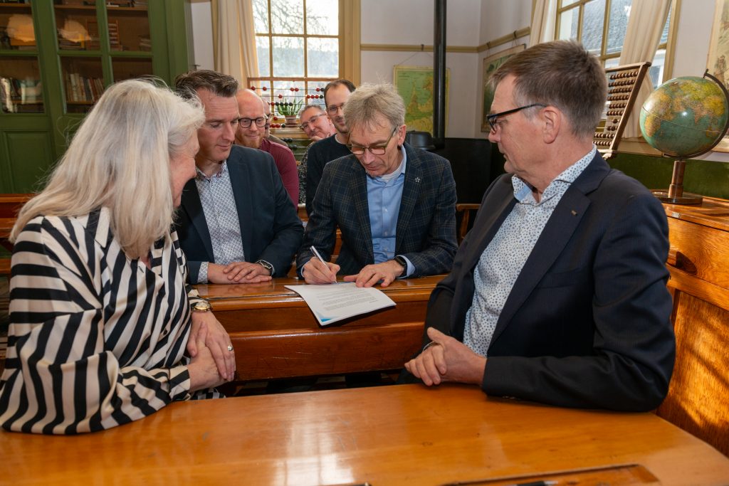 In de ouderwetse schoolbanken van het Zuiderzeemuseum zitten de acht vertegenwoordigers van de gemeente en schoolbesturen. Ze kijken hoe ze één voor één het plan ondertekenen