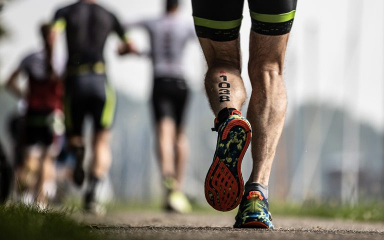 Close-up van de benen van een deelnemer aan IRONMAN Westfriesland die hardloopt over een asfaltpad, met startnummer 1038 op zijn kuit. Op de achtergrond zijn andere atleten zichtbaar, ook gekleed in sportkleding, tijdens het looponderdeel van de triatlon.