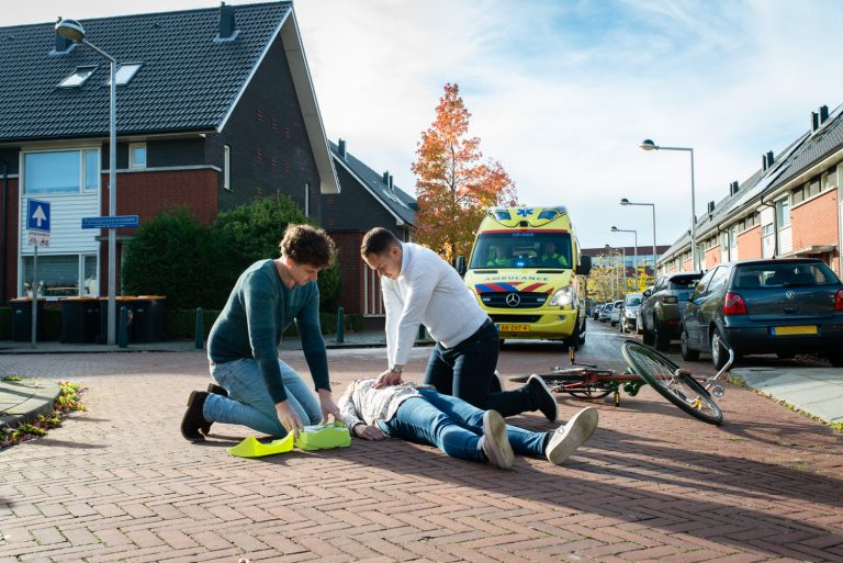 Twee personen reanimeren een derde persoon op straat. Op de achtergrond staat al een ambulance.