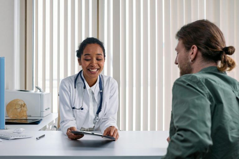 dokter in witte jas achter bureau. Bureau en raambekleding op de achtergrond is wit. Op de voorgrond een patiënt in een groen overhemd.