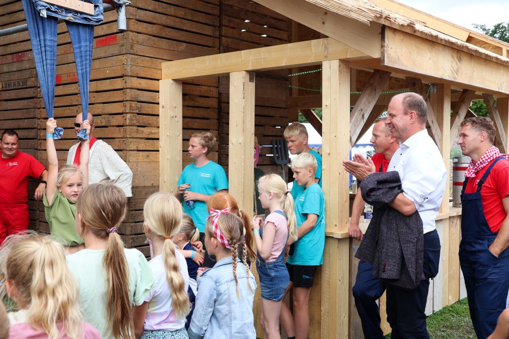 Burgemeester Bram moedigt samen met kinderen op het huttendorp een meisje aan die zo lang mogelijk probeert te hangen aan spijkerbroek.