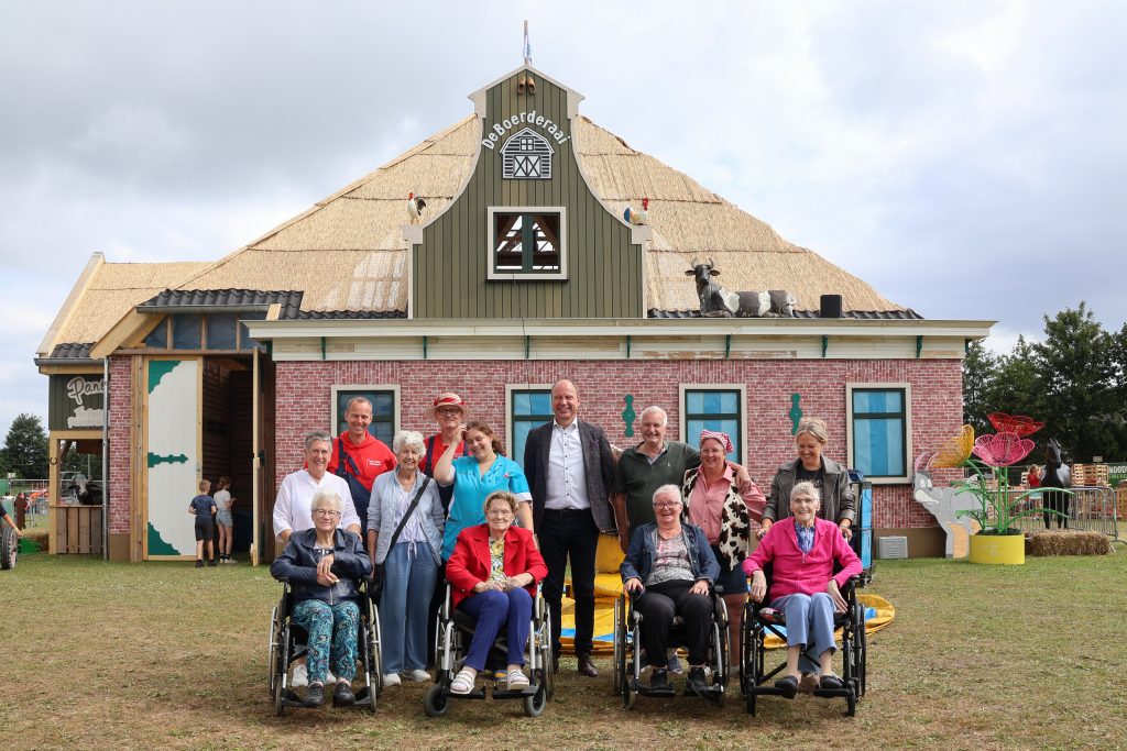 Burgemeester Bram Diepstraten samen met anderen (staand en in een rolstoel) voor de tijdelijke houten boerderij van het huttendorp.