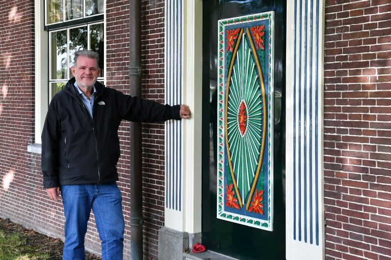 Wethouder Marcel ten Have staat voor de rouw- en trouwdeur van stolpboerderij de Wilheminahoeve. De deur is met rood en mintgroene vormen gedecoreerd.