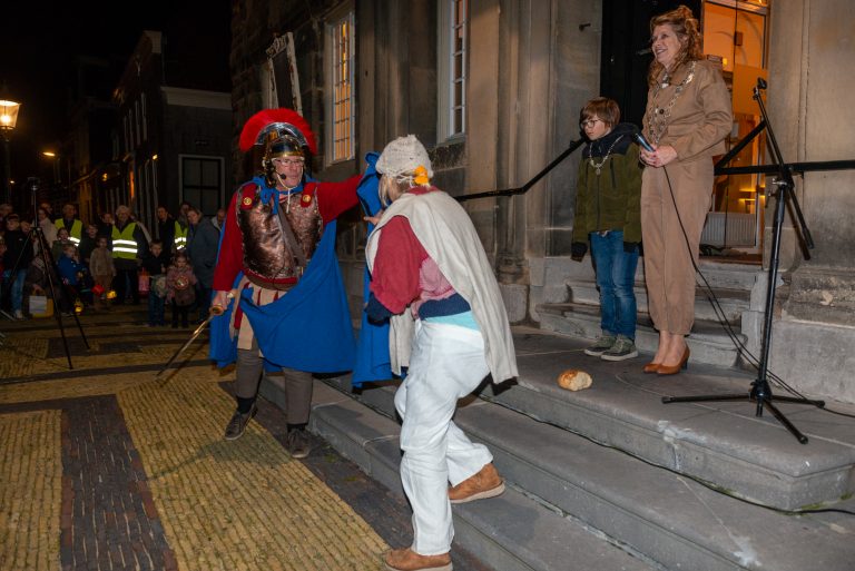 Links St. Maarten met een rode helm en blauwe mantel. Rechts de bedelaar met een witte broek en rood/witte trui. Zij neemt de mantel van St. maarten aan. Rechts staan op de trap de burgemeester en de kinderburgemeester