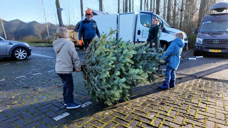 2 kinderen brengen hun kerstboom naar het inleverpunt. Een vrouw (mogelijk moeder) en man van de buitendienst kijken naar de kinderen.