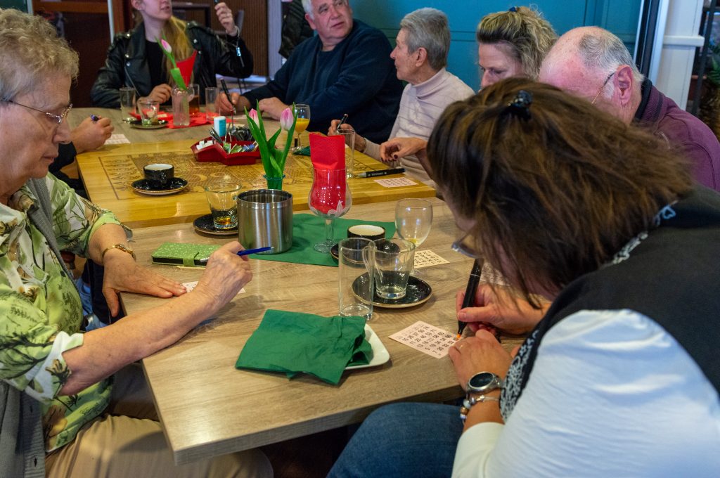 aan een lange lichtbruine tafel zitten meerdere vrouwen en mannen met een bingokaart voor hen. Rechts op de foto stempelt een vrouw haar kaart